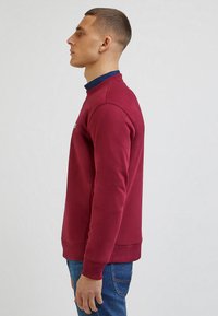 Burgundowa bluza z okrągłym dekoltem, ściągaczami przy rękawach i u dołu. Miękka tkanina z delikatnymi detalami w szwie, w zestawieniu z niebieskimi dżinsami.