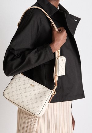 Persona con giacca nera e gonna beige plissettata porta una borsa a tracolla beige con motivo e una piccola pochette attaccata.