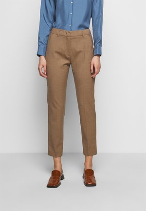 Person iført brune skræddersyede ankel-lange bukser, en blå skjorte med knapper og brune læder loafers mod en ensfarvet baggrund.