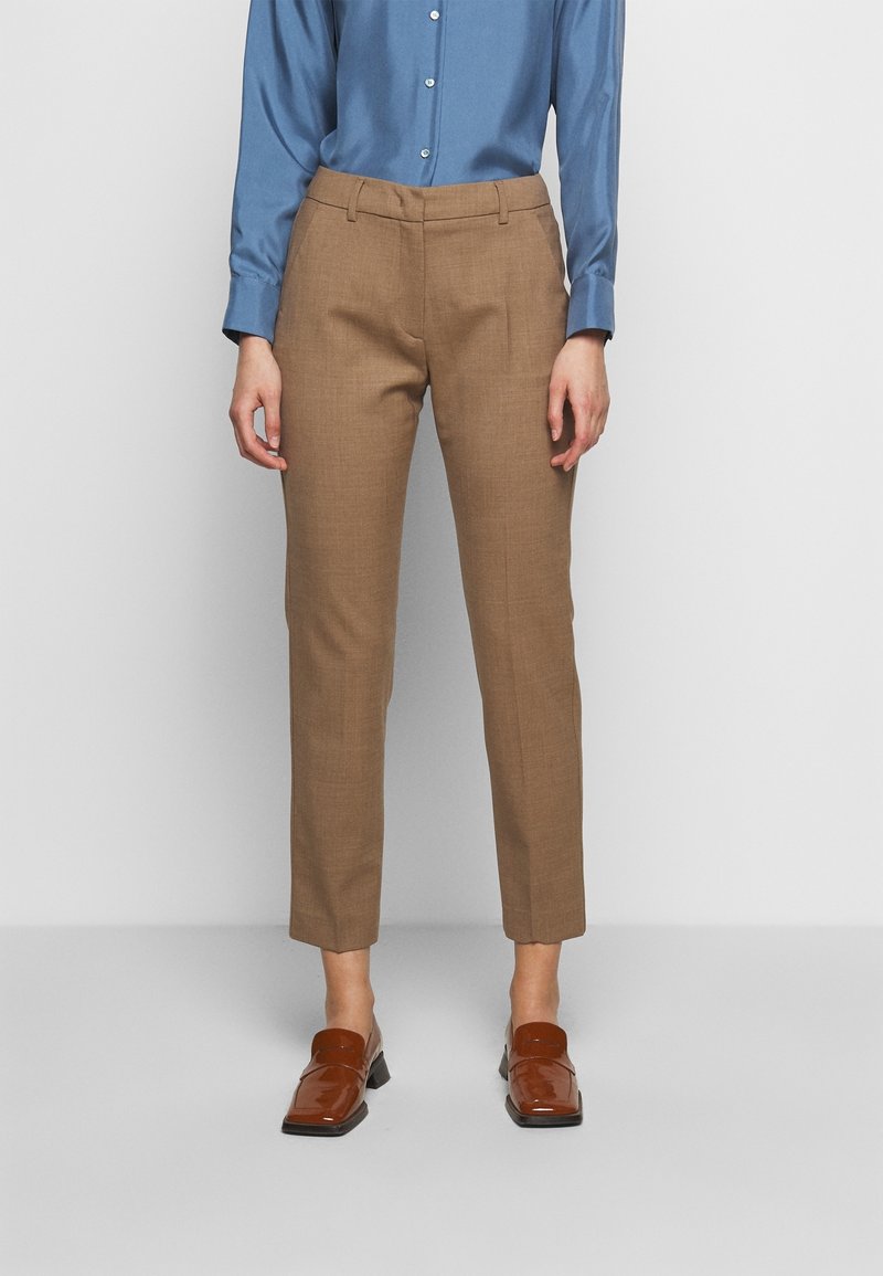 Personne portant un pantalon marron sur mesure longueur cheville, une chemise bleue boutonnée et des mocassins en cuir marron sur un fond uni.