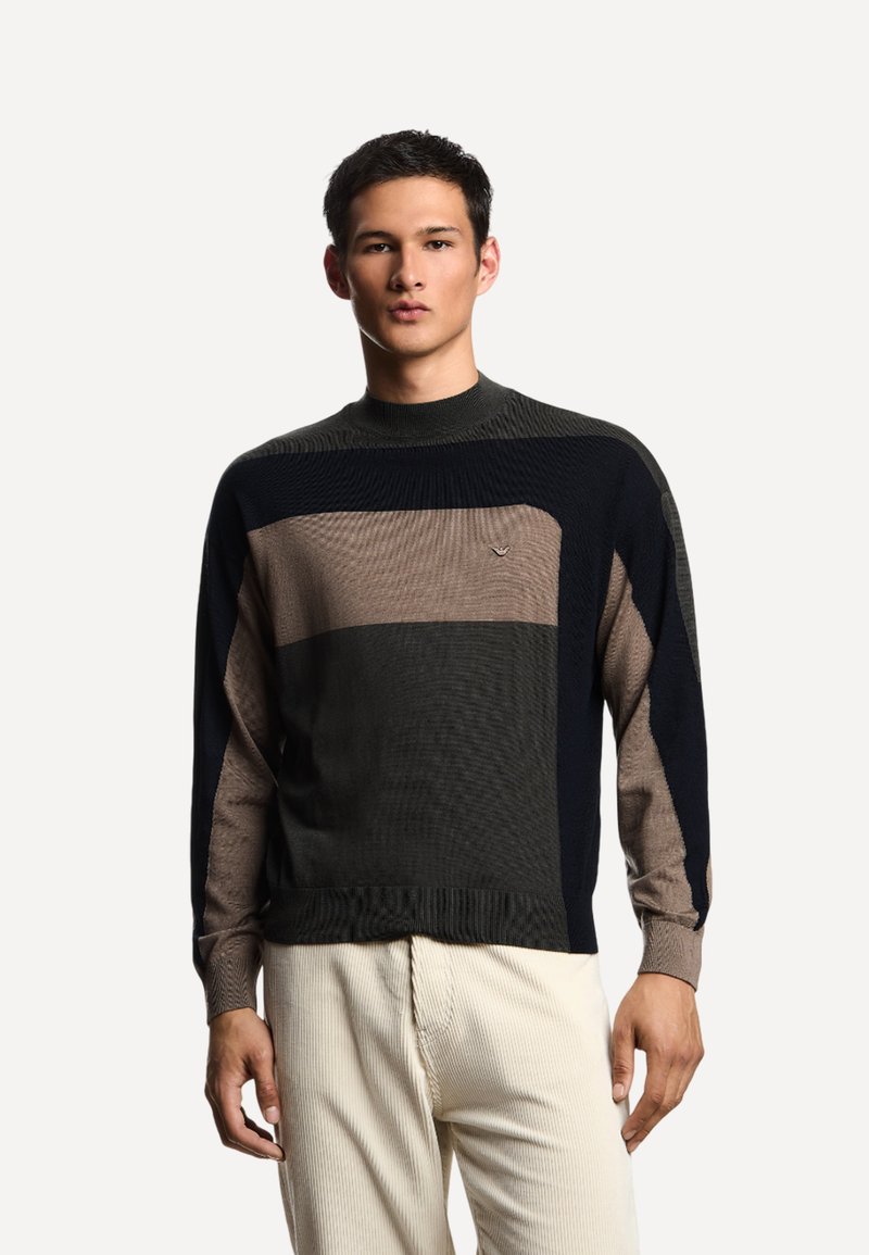 Giovane uomo che indossa un maglione a righe colorate in nero, marrone e grigio con pantaloni in velluto a coste color crema, in piedi contro uno sfondo semplice.