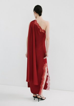 Mujer con peinado recogido en moño negro que lleva un vestido rojo de un solo hombro con ribete de encaje y abertura lateral, combinado con sandalias negras de tacón, de espaldas.