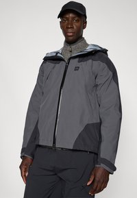 Chaqueta exterior gris con un acento negro contrastante, cierre frontal y puños ajustables. Llevada sobre un suéter gris. Cuenta con capucha.