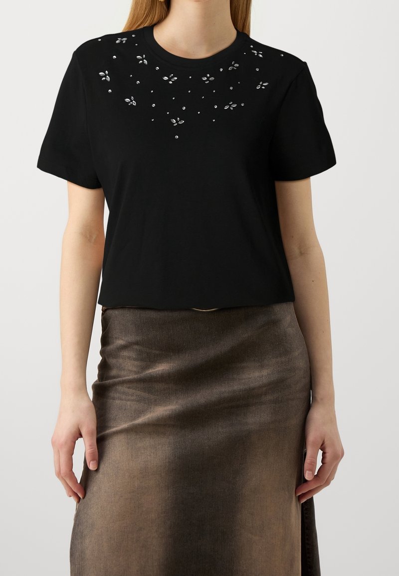 Femme portant un haut noir à manches courtes avec de petites décorations argentées près du col et une jupe midi marron brillante.