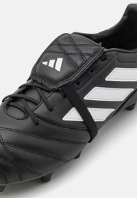 Scarpa sportiva nera con tomaia in pelle sintetica, tre strisce bianche e dettagli testurizzati. Presenta una chiusura con lacci e tacchetti in gomma.