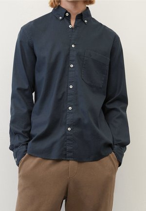 Mann trägt ein langärmeliges marineblaues Hemd mit Brusttasche und braune Hose, steht mit den Händen in den Taschen vor einem einfarbigen Hintergrund.