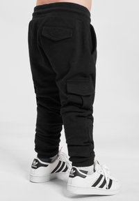 Gyerek fekete cargo jogger nadrágot és fehér Adidas Superstar sportcipőt visel fekete csíkokkal, egy sima háttér előtt áll.