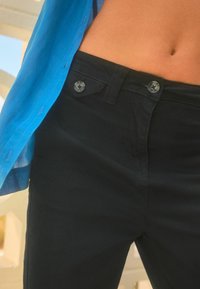 Pantaloni neri con una texture liscia, dotati di due bottini in vita e una camicia blu, parzialmente sbottonata, indossata sopra di essi.
