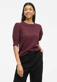 Pull bordeaux à manches courtes avec un tissu texturé, col rond et manches bouffantes, associé à un pantalon noir.