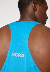 Blaues sportliches Tanktop aus atmungsaktivem, strukturiertem Stoff, mit Racerback-Design und reflektierendem HOKA-Logo auf der Rückseite.