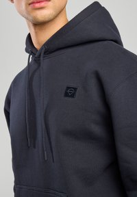 Felpa con cappuccio blu navy realizzata in tessuto, caratterizzata da un patch logo nero testurizzato sul lato sinistro del petto e da lacci regolabili con punte nere.