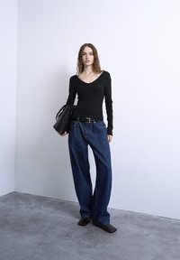 Γυναίκα με κυματιστά μαλλιά, φοράει μαύρη μπλούζα με μακριά μανίκια, φαρδιά μπλε τζιν, κρατά μαύρη τσάντα χειρός, στέκεται σε γκρι δάπεδο μπροστά σε λευκό τοίχο.