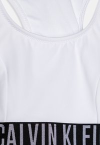 Biały sportowy top z rajstopą, charakteryzujący się fasonem racerback. U dołu znajduje się czarny pasek z nadrukiem "CALVIN KLEIN" w odstających, białych literach.