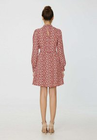 Femme portant une robe longue à manches longues à fleurs rouges avec un décolleté en forme de clé dans le dos, se tenant de dos, chaussée de sandales à talons hauts couleur chair avec une bride autour de la cheville.