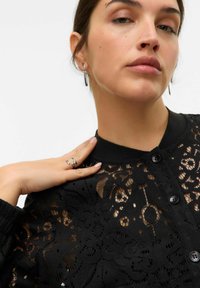 Chemisier en dentelle noire avec des motifs floraux, fermeture à boutons et col côtelé noir. Le modèle porte des boucles d'oreilles en argent et une bague, laissant apparaître sa peau en dessous.