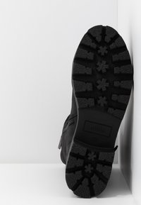 Semelle de botte noire avec des crampons profonds présentant des motifs en forme d'étoile et rectangulaires, positionnée droite contre un mur d'angle blanc.
