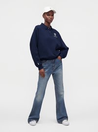Personne portant un sweat-shirt bleu marine, un jean large bleu clair, des baskets blanches et une casquette blanche, debout avec une main dans la poche.