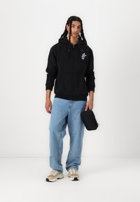 Svart hettegenser med glidelås, lys blå jeans, beige sneakers. Strukturert stoff med brodert design på brystet. Svart veske holdt i den ene hånden.