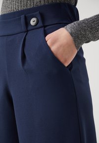 Main dans la poche d’un pantalon bleu marine à plis avec un grand bouton, associé à un haut gris côtelé à manches longues.