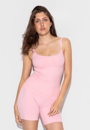 Frau mit langen braunen Haaren, die ein eng anliegendes hellrosa Tanktop und passende Shorts trägt, steht vor einem schlichten hellen Hintergrund.