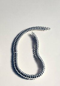 Bracelet en métal argenté brillant en forme de spirale, avec des maillons arrondis et un fermoir, posé en arc sur une surface blanche.