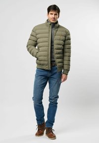 Muškarac stoji s rukom u džepu, odjeven u maslinasto zeleni puffer jaknu, sivi rolak, plave traperice i smeđe kožne čizme na bijeloj pozadini.