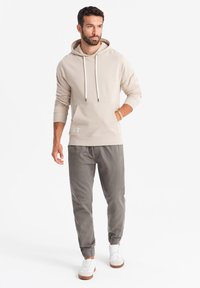Hellbeige Kapuzenpullover mit Kordelzug und Fronttasche, kombiniert mit grauen schmal geschnittenen Hosen und weißen Sneakers mit sandfarbenen Sohlen.