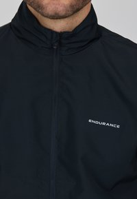 Mörkblå vindjacka med hög krage och framdragkedja. Har en vit "ENDURANCE"-logotyp på vänster sida. Slät, lättviktig tyg.