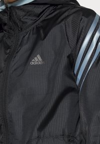 Veste noire Adidas avec un motif quadrillé discret, trois bandes bleu clair sur les manches et une fermeture éclair devant avec le logo Adidas sur la poitrine.