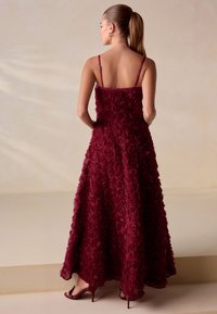 Bordeauxrode jurk met een getextureerd rozenpatroon, spaghettibandjes en een vloeiende silhouet. Gecombineerd met sandalen met hoge hakken en bandjes.