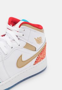 Bílé tenisky s zlatými akcenty, červenými a modrými detaily a texturovanou červenou vzorovanou patou. Obsahují bílé tkaničky a logo Air Jordan.