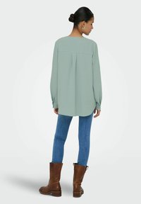 Femme aux cheveux relevés en chignon, portant des boucles d'oreilles créoles argentées, un chemisier ample à manches longues vert clair, un jean bleu et des bottes marron mi-mollet, debout, tournée vers l'arrière.