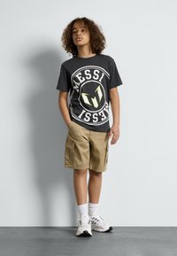 Svart t-skjorte med hvit og neon grafikk som viser "MESSI" og logo. Beige cargo shorts, hvite sokker og svarte joggesko med detaljer.