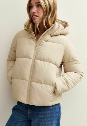 Beige pufferjack met een capuchon, gequilt textuur, voorkant met rits en zijzakken, gecombineerd met blauwe spijkerbroek.
