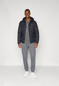 Black puffer jacket with a hood, gray sweatshirt underneath, gray sweatpants, and white sneakers. Model stands against a neutral background.