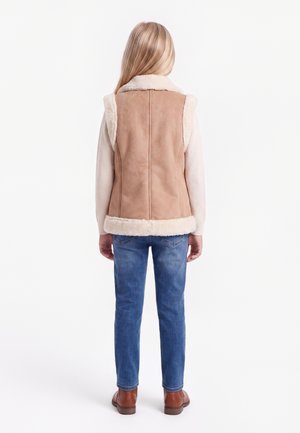 Barn med langt blond hår iført tanfarvet ruskindsvest, hvid langærmet skjorte, blå jeans og brune støvler, står og vender væk.