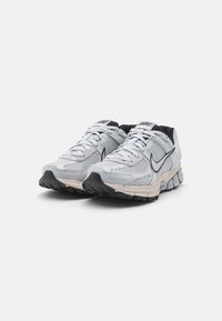 Grå mesh löparskor med svarta detaljer, rundad tå, vadderad sula och framträdande snörningssystem. Logotypen visas på sidan.