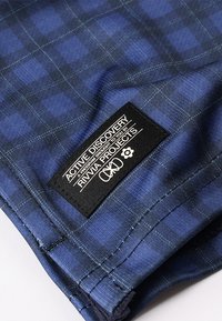 Kék kockás szövet sima textúrával; az alsó szélén egy fekete címke van varrva, amelyen fehér szöveg és logók találhatók.