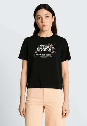 Kobieta z długimi ciemnymi włosami, ubrana w czarną koszulkę z kwiatowym nadrukiem i napisem „Design Studio Denim Tom Tailor” oraz beżowe spodnie.