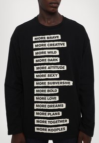 T-shirt nero a maniche lunghe con testo bianco audace stampato in formato elenco, con un effetto testurizzato simile alla carta sulle lettere.