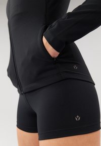 Schwarze Sportjacke mit Reißverschluss vorne, strukturiertem Stoff und Seitentaschen, kombiniert mit schwarzen, figurbetonten Shorts, die ein dezentes Logo zeigen.