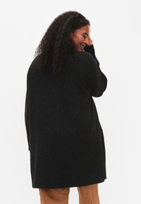 Schwarze Strickjacke mit lockerer Passform, langen Ärmeln und Seitentaschen. Die Textur wirkt weich mit einem leichten Glanz. Hoch-Tief-Saum.