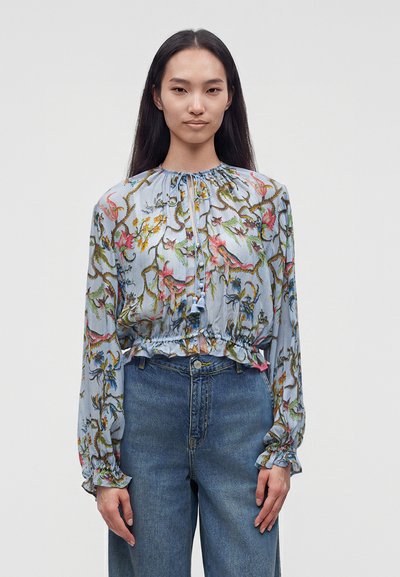 Femme aux longs cheveux foncés portant un chemisier bleu clair à fleurs à manches longues et un jean bleu taille haute, debout devant un fond uni.