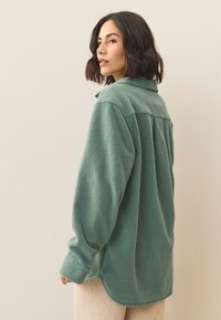 Maglia in pile verde spento, con scollo con colletto, maniche lunghe e vestibilità comoda con orlo leggermente curvato. Tessuto strutturato.