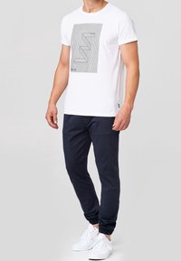Biała bawełniana koszulka z czarnym wzorem graficznym, zestawiona z granatowymi spodniami stretch. Model nosi białe sneakersy, prezentując swobodny strój.