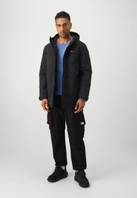 Man som står och bär en svart huva jacka, blå skjorta, svarta cargobyxor och svarta sneakers mot en enkel ljus bakgrund.