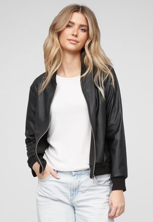 Blonde Frau trägt eine schwarze Lederjacke über einem weißen Oberteil und hellblauen Jeans, steht mit den Händen in den Taschen vor grauem Hintergrund.