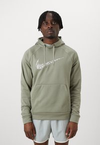 Zelená mikina s kapucí vyrobená z měkké látky, s velkým bílým logem Nike na hrudi, klokankovou kapsou a stahovací kapucí.