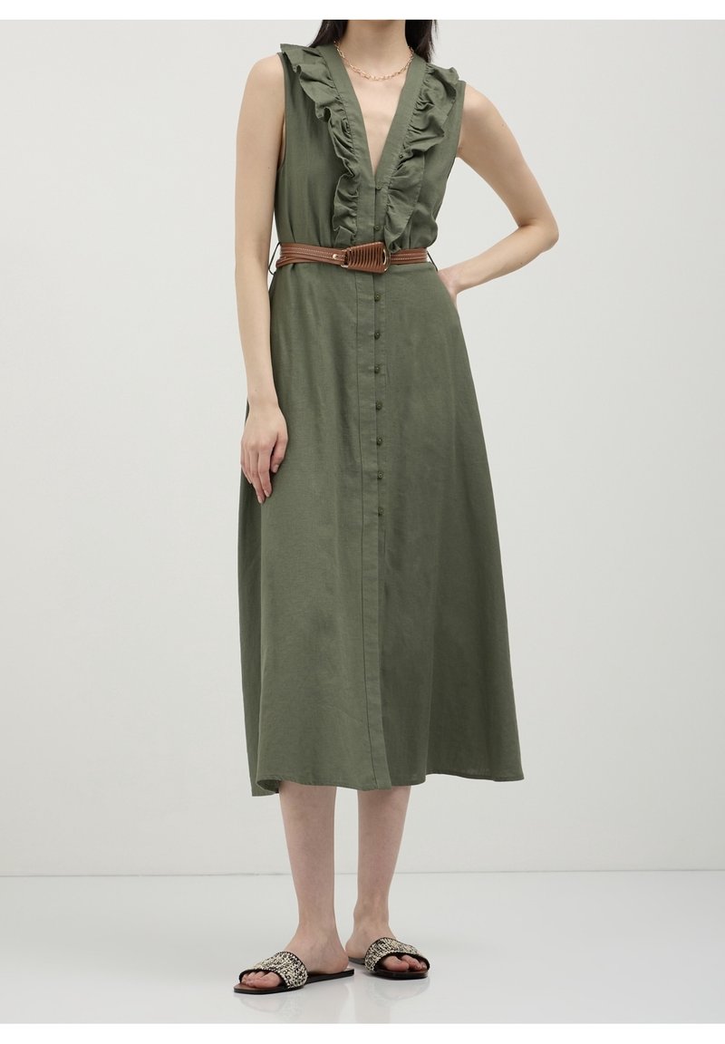 Robe midi sans manches vert olive avec décolleté en V volanté, boutonnée devant, ceinture marron, et sandales plates à motifs sur une personne debout.