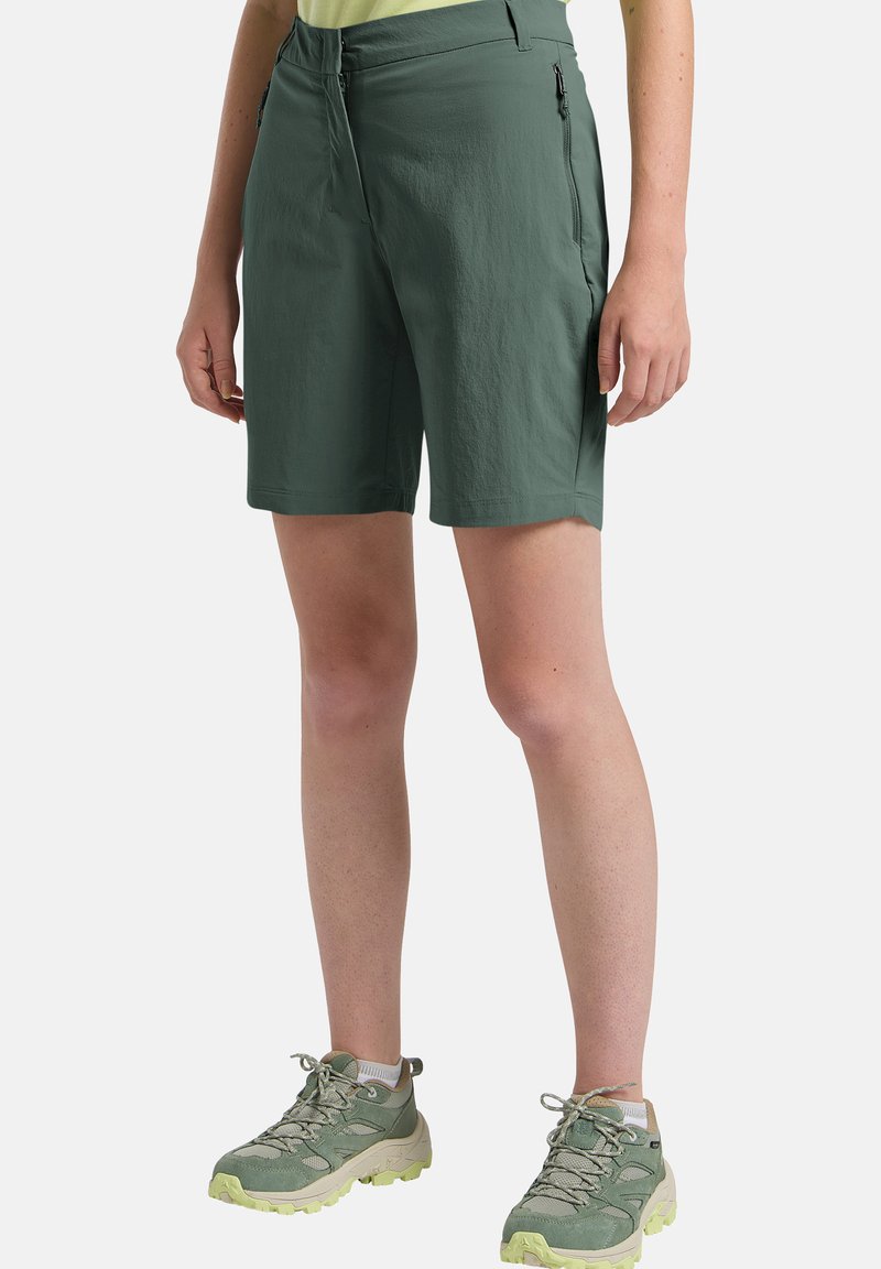 Jambes portant un short de randonnée vert jusqu'aux genoux avec des poches zippées et des chaussures de randonnée vertes à semelles épaisses.